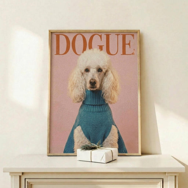 May include: Framed poster showing a white poodle wearing a teal blue turtleneck sweater against a soft pink background, with "DOGUE" text in orange letters at top. Gold frame sits on light wood shelf next to small wrapped white gift box with evergreen sprig. Clean white wall background with natural lighting creates shadow on wall.