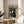 May include: Black and white vintage Japanese poster artwork featuring stylized cats in a geometric dotted pattern, displayed in a thin wooden frame on a round wooden side table next to a beige upholstered chair. Natural sunlight streams across the neutral-toned room setting with plant leaves visible in the corner.
