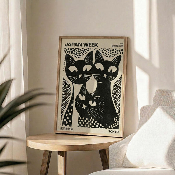 May include: Black and white vintage Japanese poster artwork featuring stylized cats in a geometric dotted pattern, displayed in a thin wooden frame on a round wooden side table next to a beige upholstered chair. Natural sunlight streams across the neutral-toned room setting with plant leaves visible in the corner.