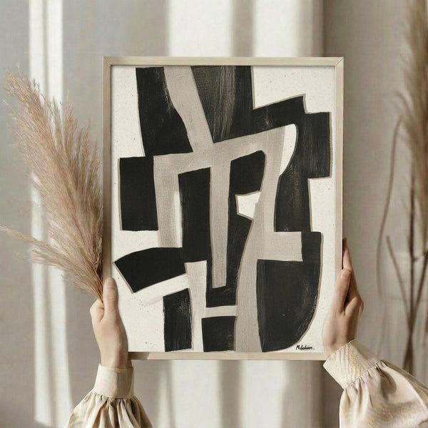 May include: Hands holding a framed abstract artwork featuring bold black brushstrokes on cream background, displayed next to dried pampas grass stems. The geometric composition shows intersecting angular shapes with natural brush textures. Light wood frame complements the neutral beige clothing and soft window lighting in the styled interior setting.