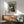 May include: Framed black and white vintage poster featuring three stylized cats with closed eyes arranged in a group, surrounded by dotted patterns. The artwork displays "JAPAN WEEK" and "TOKYO" text with Japanese characters. Shown hanging above a wooden sideboard with sliding doors and ceramic table lamp, next to a mid-century modern green upholstered armchair with wooden frame. Natural sunlight creates shadows on white wall.