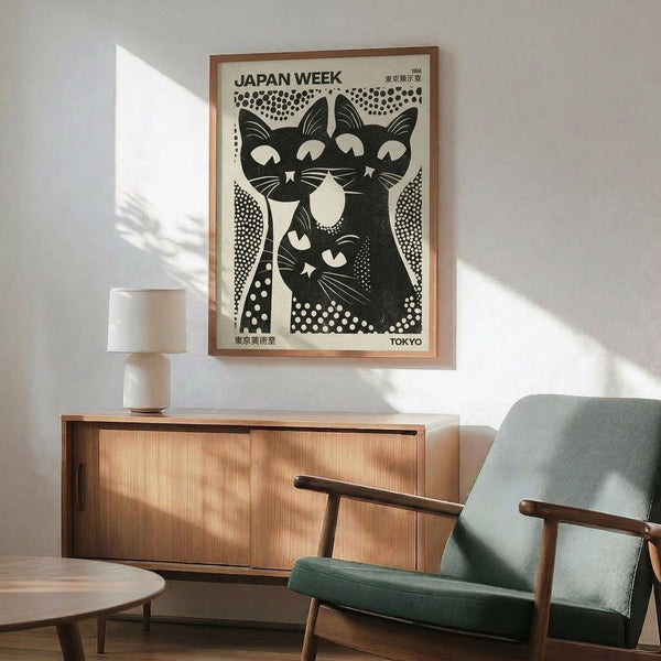 May include: Framed black and white vintage poster featuring three stylized cats with closed eyes arranged in a group, surrounded by dotted patterns. The artwork displays "JAPAN WEEK" and "TOKYO" text with Japanese characters. Shown hanging above a wooden sideboard with sliding doors and ceramic table lamp, next to a mid-century modern green upholstered armchair with wooden frame. Natural sunlight creates shadows on white wall.