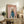 May include: Framed poster featuring a white poodle wearing a blue turtleneck sweater against a pink background with "DOGUE" text in orange letters, displayed on a rustic wooden sideboard alongside books, a ceramic vase, and a fern plant, with a beige armchair visible in a cozy living room setting.
