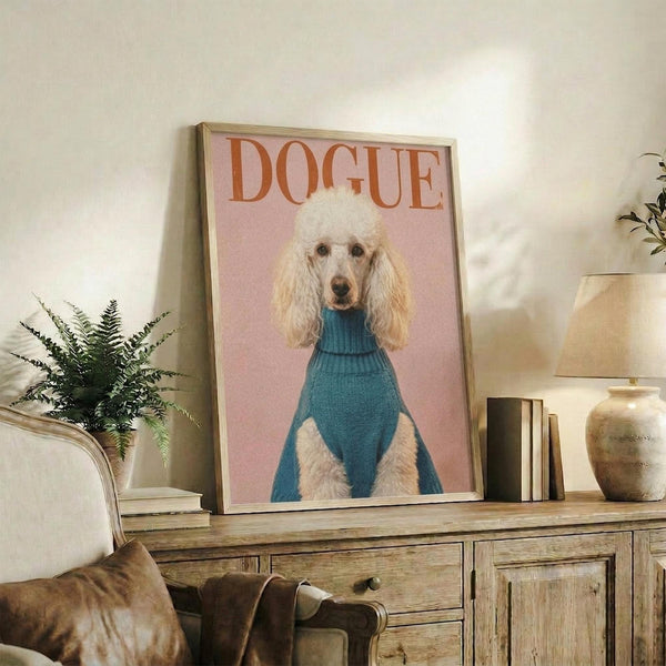 May include: Framed poster featuring a white poodle wearing a blue turtleneck sweater against a pink background with "DOGUE" text in orange letters, displayed on a rustic wooden sideboard alongside books, a ceramic vase, and a fern plant, with a beige armchair visible in a cozy living room setting.