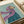 May include: Whimsical painted portrait of a tan Italian Greyhound wearing a flowing purple headscarf and teal coat against a soft mint green background. The artwork is displayed in a light wood frame on white wrapping paper with cream ribbon nearby. The painting has visible brushstrokes and captures the dog's gentle expression and elegant pose in an folk art style.