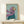 May include: Framed painting of an Italian Greyhound wearing a flowing purple headscarf and teal coat against a soft mint green background, displayed on a light wood shelf next to a small wrapped gift with twine. The artwork has visible brushstroke texture and is housed in a thin natural wood frame.