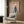 May include: Framed poster print on wood-paneled wall above beige upholstered armchair with geometric pillow. Artwork shows white poodle wearing teal blue turtleneck sweater against soft pink background with "DOGUE" text at top. Modern living room setting with brass floor lamp, side table with open book, and natural lighting from window with curtains.