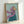 May include: Framed painting of a tan Italian Greyhound wearing a flowing purple headscarf and teal garment against a mint green background. The artwork shows visible brushstrokes and is displayed in a light wooden frame, leaning against a white wall. The dog has large dark eyes and is positioned in a three-quarter view portrait style.