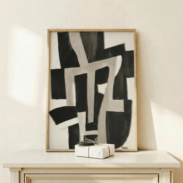 May include: Black and white abstract geometric artwork in light wood frame, displayed on pale wooden console table against cream wall. Small wrapped gift box with greenery sits nearby. Bold brushstroke patterns create interlocking angular shapes across white canvas background.