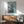 May include: Framed artwork showing an Italian Greyhound wearing a purple headscarf and teal garment against a mint green background, displayed above a wooden sideboard with lamp in a mid-century modern living room with gray upholstered chair and natural lighting casting geometric shadows on white walls.