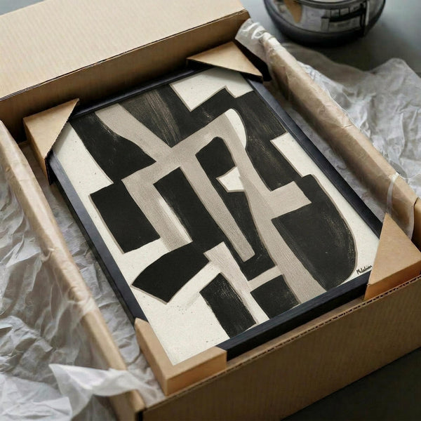 Abstract geometric artwork with bold black angular shapes on cream background, displayed in black frame inside cardboard shipping box with protective wrapping and wooden handle tools visible alongside