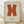 May include: Framed poster with large orange X shape on cream background, displayed in light wood frame against white wall on wooden floor with natural sunlight casting shadows