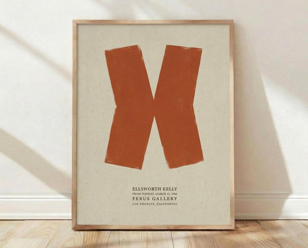 May include: Framed poster with large orange X shape on cream background, displayed in light wood frame against white wall on wooden floor with natural sunlight casting shadows