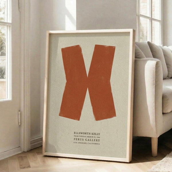 May include: Large framed poster with orange X shape on cream background, displayed in bright living room with beige sofa, white walls, and tall windows. Poster shows exhibition text for Ellsworth Kelly at Ferus Gallery Los Angeles. Light wooden frame sits on hardwood floor against window, creating warm natural lighting and shadows.