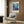 May include: Framed underwater scene poster with colorful coral reef and tropical fish in blues, purples, and oranges, displayed above a beige upholstered armchair with geometric patterned pillow. Modern brass floor lamp and dark side table with open book complete the contemporary living room setting against wood-paneled wall.