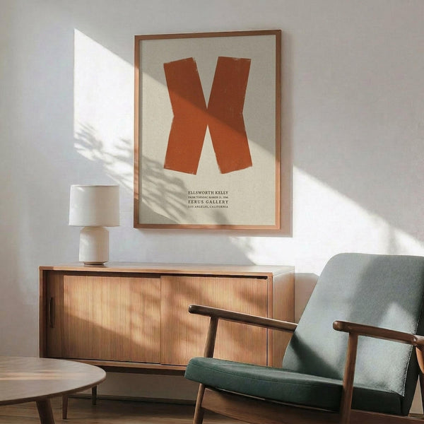 May include: Framed vintage art poster featuring a bold orange X shape on cream background with gallery text below, displayed above a wooden sideboard with white lamp, next to a sage green mid-century modern armchair. Warm sunlight creates dramatic shadows across the white wall and furniture in this styled living room setting.