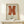 May include: Orange geometric abstract artwork in wooden frame displayed on light wood shelf against cream wall. Modern minimalist poster features bold rust-colored angular shapes on beige background with black text below. Small white wrapped gift with twine and pine sprig sits beside frame. Warm natural lighting creates soft shadows.