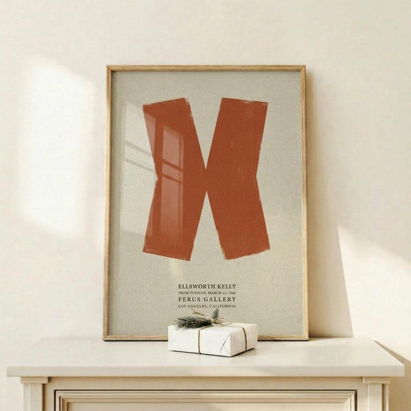 May include: Orange geometric abstract artwork in wooden frame displayed on light wood shelf against cream wall. Modern minimalist poster features bold rust-colored angular shapes on beige background with black text below. Small white wrapped gift with twine and pine sprig sits beside frame. Warm natural lighting creates soft shadows.
