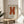 May include: Orange letter X poster in thin wood frame displayed on round wooden stool next to beige upholstered chair. Sunlight streams across white wall creating soft shadows. Green plant leaves visible in corner. Minimalist modern home decor setting with neutral tones and natural textures.