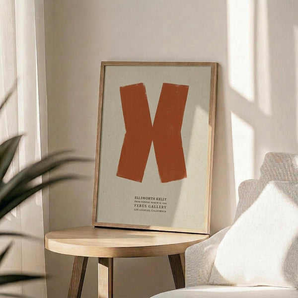 May include: Orange letter X poster in thin wood frame displayed on round wooden stool next to beige upholstered chair. Sunlight streams across white wall creating soft shadows. Green plant leaves visible in corner. Minimalist modern home decor setting with neutral tones and natural textures.
