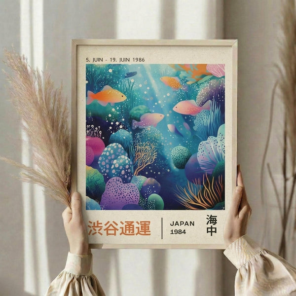 May include: Person holding framed vintage-style poster featuring colorful underwater scene with tropical fish, coral, and sea plants in vibrant blues, purples, oranges and pinks. Japanese text and "JAPAN 1984" visible at bottom. Natural wood frame, dried pampas grass visible, soft beige clothing and neutral background.