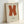 Framed minimalist art print featuring a large rust-orange letter K on cream textured paper background. The geometric letterform has clean, bold lines with slightly irregular painted edges. Below the K is small black text reading "Ellsworth Kelly" and exhibition details. The print is displayed in a thin natural wood frame, leaning against a white wall on what appears to be a light wooden surface. Soft natural lighting creates gentle shadows around the frame.