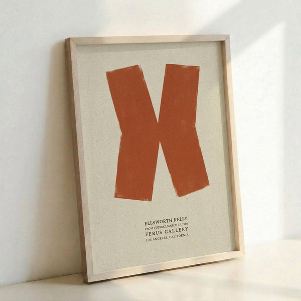 Framed minimalist art print featuring a large rust-orange letter K on cream textured paper background. The geometric letterform has clean, bold lines with slightly irregular painted edges. Below the K is small black text reading "Ellsworth Kelly" and exhibition details. The print is displayed in a thin natural wood frame, leaning against a white wall on what appears to be a light wooden surface. Soft natural lighting creates gentle shadows around the frame.