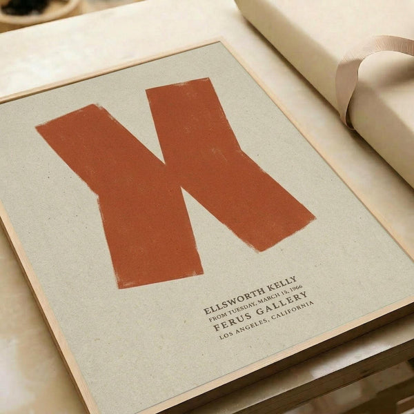 May include: Orange letter "N" poster print displayed on cream colored paper with black text reading "Ellsworth Kelly" and "Ferus Gallery" below, shown alongside rolled paper tubes on a neutral surface in warm lighting