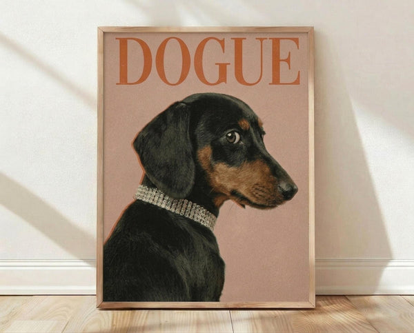 May include: Framed poster print featuring a black and tan dachshund wearing a sparkly rhinestone collar against a dusty pink background, with "DOGUE" in large orange serif letters across the top. The wooden frame sits on light hardwood flooring against a white wall with soft natural lighting creating diagonal shadows.