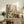 May include: Framed botanical art print leaning against white wall on rustic wooden sideboard, featuring stylized wildflowers and plants in purple, yellow, green, and brown dotted patterns. Scene shows fern plant, ceramic vase, books, and brown leather chair arm, creating cozy living room display with warm natural lighting.
