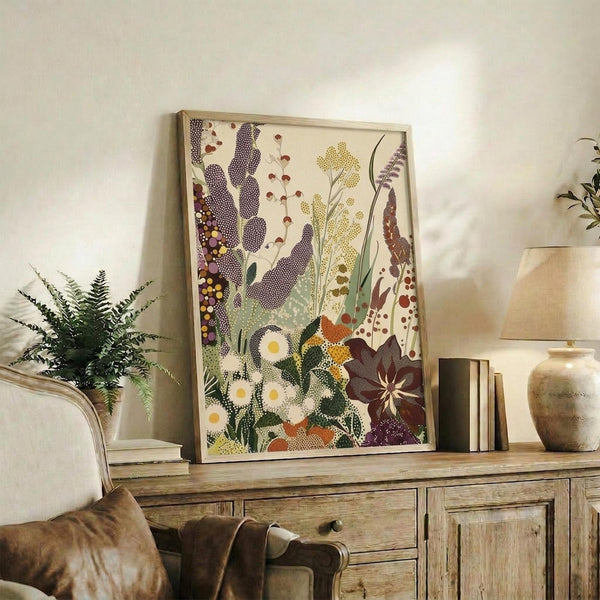 May include: Framed botanical art print leaning against white wall on rustic wooden sideboard, featuring stylized wildflowers and plants in purple, yellow, green, and brown dotted patterns. Scene shows fern plant, ceramic vase, books, and brown leather chair arm, creating cozy living room display with warm natural lighting.
