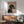 May include: Framed poster showing a black and tan dachshund in profile wearing a rhinestone collar against a peachy pink background, with "DOGUE" text at top in fashion magazine style. Displayed on wooden sideboard next to white table lamp, with mint green mid-century modern chair partially visible. Warm sunlight streams across white wall creating geometric shadows.
