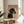 May include: Framed poster featuring a black and tan dachshund wearing a pearl necklace against a peachy pink background with "DOGUE" text in orange serif letters, displayed on a light wood round side table next to a beige upholstered chair with white pillow, sunlight streaming across white walls with plant shadows visible