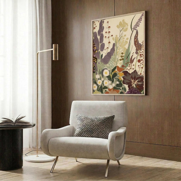 May include: Modern beige armchair with geometric pillow positioned against wood-paneled wall, featuring botanical art print with colorful wildflowers, seed pods, and meadow plants in purple, yellow, green, and brown tones. Gold floor lamp and dark round side table with open book complete the contemporary room setting with natural lighting from window.