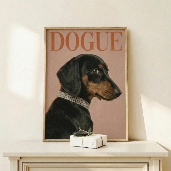 May include: Framed poster featuring a black and tan dachshund wearing a rhinestone collar on dusty pink background with "DOGUE" text in orange letters, displayed on light wood surface next to small white wrapped gift with twine bow against cream wall