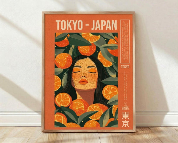 May include: Framed poster with orange background featuring stylized woman with black hair surrounded by oranges, orange slices, and green leaves. Text reads "TOKYO - JAPAN" at top with Japanese characters at bottom. Wooden frame sits on light wood floor against white wall with fabric shadows visible.