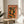 May include: Framed orange and green poster featuring an illustrated woman with closed eyes surrounded by oranges and citrus slices, displayed on a round wooden side table next to a beige upholstered chair in a bright room with white walls and natural sunlight casting shadows from a plant.