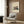 May include: Cream-colored poster with modernist architectural building illustration featuring geometric shapes in teal, orange, and beige, displayed in light wood frame on wood-paneled wall above beige upholstered armchair with geometric patterned pillow, brass floor lamp and dark round side table with open book nearby