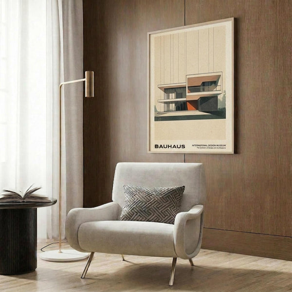 May include: Cream-colored poster with modernist architectural building illustration featuring geometric shapes in teal, orange, and beige, displayed in light wood frame on wood-paneled wall above beige upholstered armchair with geometric patterned pillow, brass floor lamp and dark round side table with open book nearby