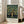 May include: Framed poster featuring William Morris-style apple tree design with golden fruit and green foliage on dark background, exhibition text at bottom reading "Centenary Exhibition William Morris 1934 Textile Industry London Jan 6 to May 6 Free Admission." Light wood frame leaning against white wall next to beige upholstered sofa in bright room with large windows.