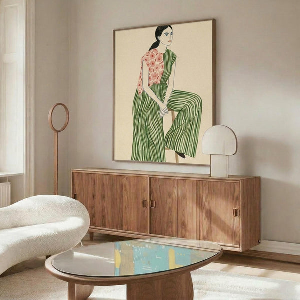 May include: Framed art print of a woman in green striped wide-leg pants and floral pink top, sitting against cream background. Displayed above mid-century walnut credenza with brass handles. Modern white curved chair and glass-top oval coffee table with abstract blue artwork underneath. Brass floor lamp and table lamp complete the styled living room setting.