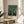 May include: Framed poster featuring a decorative tree with golden apples and green foliage on dark teal background, displayed on round wooden side table next to beige upholstered chair. Natural sunlight streams across white wall, creating soft shadows. Plant leaves visible in corner. Exhibition-style poster with white text band at bottom in light wood frame.