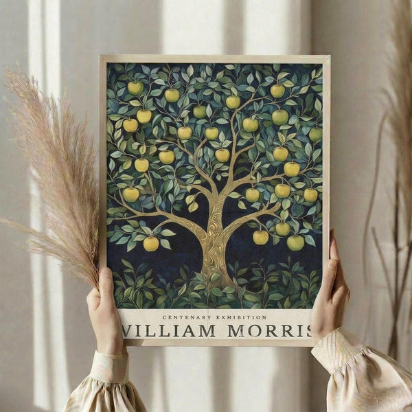 May include: Hands holding a framed William Morris centenary exhibition poster featuring an ornate apple tree with golden yellow fruit against a dark blue background. The decorative tree has intricate golden branches and lush green foliage in Morris's signature botanical style. Person wears a cream-colored sweater with gathered sleeves and holds dried wheat stems. Soft natural lighting on neutral curtained background.