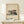 May include: Framed modernist architectural poster showing geometric concrete building with cantilevers in beige, orange and dark green tones, displayed on light wood surface beside white wrapped gift with pine sprig against cream wall