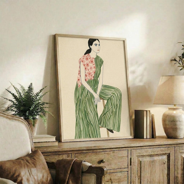 May include: Framed art print featuring a seated woman with black hair wearing a pink floral top and green striped wide-leg pants against cream background. Light wood frame displayed on rustic wooden sideboard alongside fern plant, ceramic vase, books, and brown leather cushions in neutral-toned living room setting.