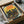 May include: Framed vintage-style poster featuring three black cats with bright yellow and green eyes against autumn sunset sky. Red "JAPAN WEEK" text at top, orange maple branches in foreground, silhouetted bird in flight. Warm yellow, orange and teal color palette with retro travel poster aesthetic. Frame packed in cardboard shipping box with protective paper wrapping on white tissue paper background.