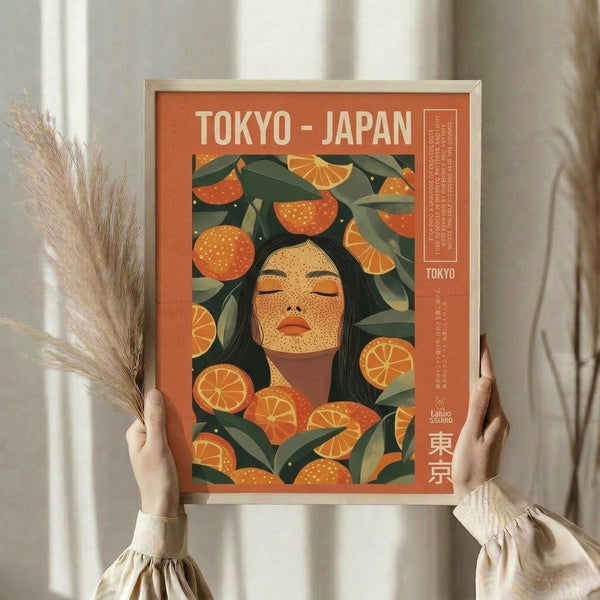 May include: Framed orange and green art print held by person in cream clothing, featuring stylized woman with closed eyes surrounded by citrus fruits and leaves, displayed near dried pampas grass against white curtained window
