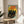 May include: Framed poster print featuring three black cats with bright green and yellow eyes against a yellow sun and turquoise sky background, with red "JAPAN WEEK" text and autumn branch details. Print sits on light wood round side table next to beige upholstered chair in bright, naturally-lit room with white walls and plant shadows.