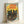 May include: A framed vintage-style travel poster featuring two black cats with bright yellow-green eyes against a sunset backdrop. Red text reads "JAPAN WEEK" at the top, with orange cherry blossoms in the foreground, flying birds, and Japanese characters. The artwork has warm yellow, orange and teal tones in a weathered, retro aesthetic. Light wooden frame sits against a white wall corner.