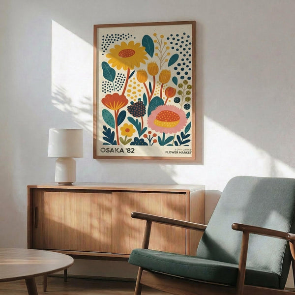 May include: Colorful floral poster with stylized flowers, leaves, and dotted patterns in yellow, orange, pink, and teal, displayed in wooden frame above mid-century modern credenza. Scene shows green upholstered armchair with wood arms, white table lamp, and round coffee table in bright, naturally-lit room with white walls.